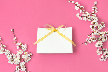 White gift bag with gold ribbon and branch of spring white flowers on bright pink background. Greeting card with delicate flowers Pink floral background. Spring minimal concept. Flat lay top view