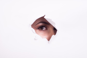 eyes of a young curious woman peeking through a hole torn in white paper poster. with beautiful make-up.