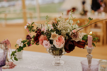 Burgundy, pink and white flowers centerpiece, white lace table linen, burning candles. Elegant wedding, event, dine dinning decor in marquee, outdoor.