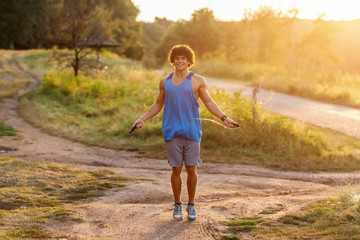 Smiling Caucasian man with curly hair and in blue t-shirt skipping rope in nature on sunny summer day. Full length.