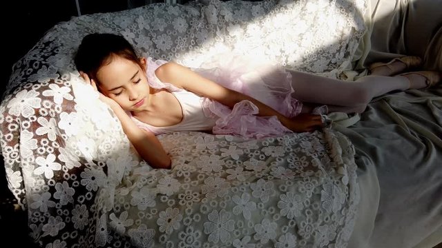 4K selective focus stabilize medium shot of beautiful ballerina girl in pink leotard and tutu sleeping on sofa cover with white lace fabric. Cute little girl in ballet dress and shoe sleeping.