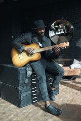 Musician in black trench coat and bucket hat sitting on bed and playing guitar. Songwriter in loft...