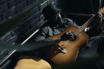 Musician in black trench coat and bucket hat sitting on bed and playing guitar. Songwriter in loft...
