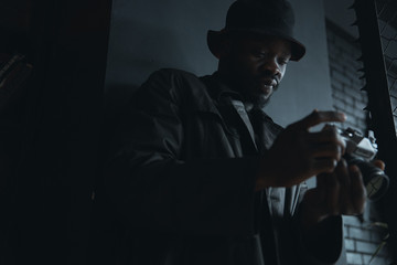 Dark portrait of young fashionable man in black trench coat and bucket hat, with camera in hands.