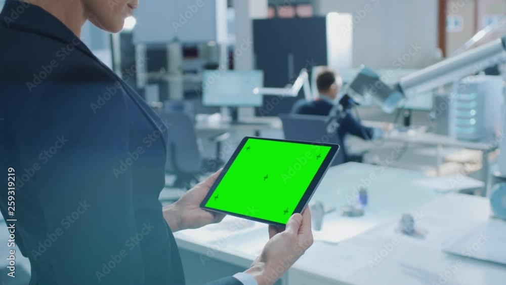 Poster Engineer Holds Green Mock-up Screen Digital Tablet Computer. In the Background Team of Industrial Engineers Working with Functional Robot Arm in Modern Bright Research Facility