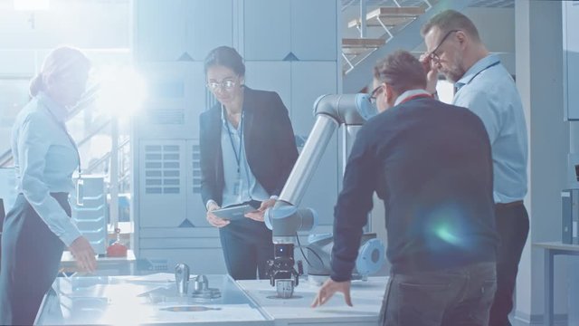 Diverse Team of Industrial Robotics Engineers Gathered Around Table With Robot Arm, They Manipulate and Program it to Pick Up and Move Metal Component.
