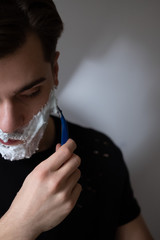Man shaving himself close-up. Using shaving cream and disposable razor. Looking to a mirror. Morning routine. Huge beard.