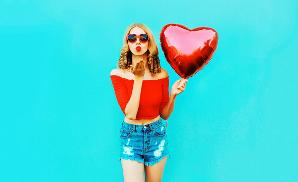Portrait beautiful young woman sending sweet air kiss with red heart shaped balloon on colorful blue background