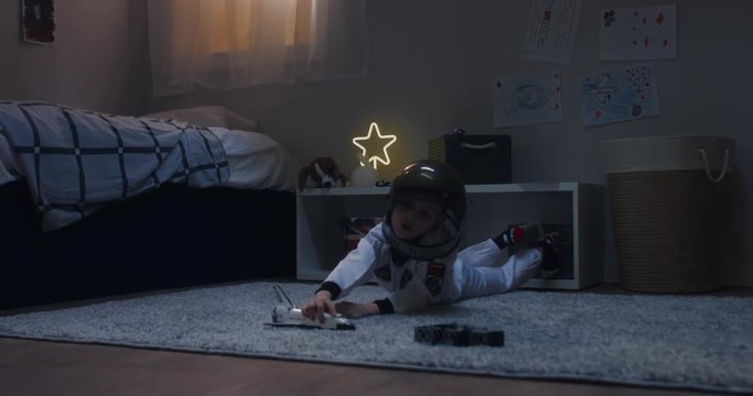 Cute little kid boy wearing astronaut suit playing with toy shuttle space ship rocket at home, bedroom interior evening shot. 4K UHD RAW FOOTAGE