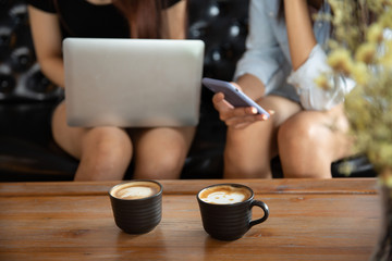 Two young girl friends with smartphone on hand talking and drinking sitting on a sofa in the coffee cafe.