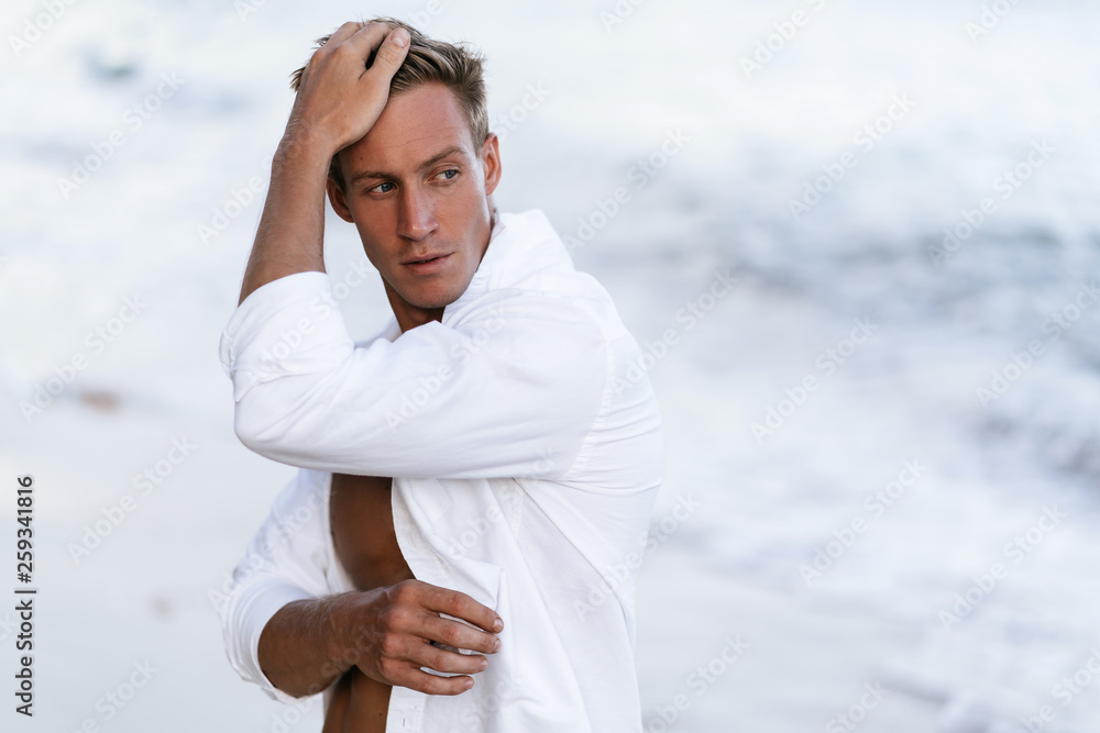 Wall mural Portrait of handsome sexy man in white shirt posing on beach, ocean waves at background