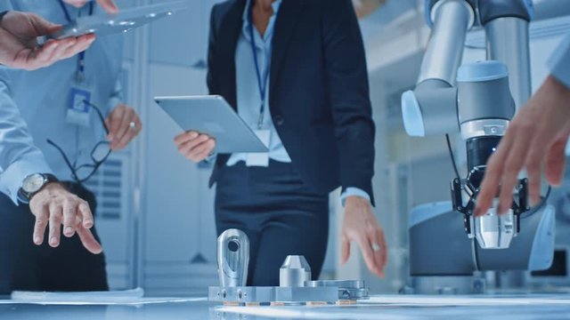 Team of Industrial Robotics Engineers Gathered Around Table With Robot Arm, They Use Tablet Computer to Manipulate and Program it to Pick Up and Move Metal Component. Focus on Hands
