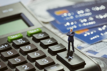 Miniature businessman standing on black calculator reflect with sun light on pile of credit cards and US Dollar bills money using as debt crisis, financial calculation or accounting concept