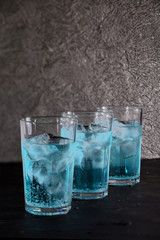 Three glasses of a cocktail based on soda and blue Curacao liqueur with ice stand in a row on a dark textured background. Preparation of cocktails.