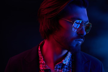 Close up neon light portrait of handsome male model with dark medium length hair, mustaches and beard in sunglasses, weared in dark jacket and floral shirt.