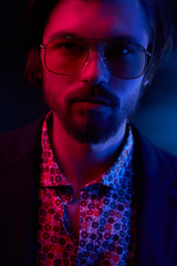 Close up neon light portrait of handsome male model with dark medium length hair, mustaches and beard in sunglasses, weared in dark jacket and floral shirt.