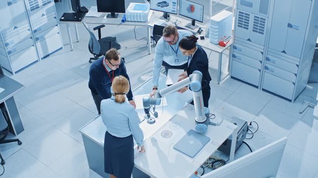 Team of Industrial Robotics Engineers Gathered Around Table With Robot Arm, They Use Tablet Computer to Manipulate and Program it to Pick Up and Move Metal Component