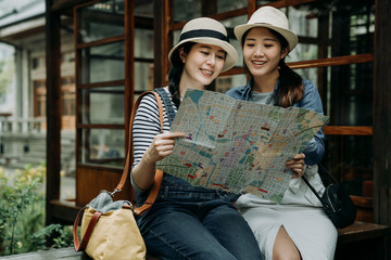 two female asian tourist with paper map sitting in bench by japanese style garden wooden house....