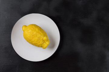 One lemon on a white plate on a dark natural colored background in Horizontal orientation. Trendy ugly organic fruit. Copy space.