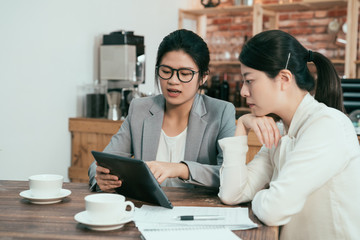 female graphic designer pointing with finger on tablet pc during collaboration with office colleague on common project in coffee shop. Two businesswomen discussing website on digital pad in cafe.