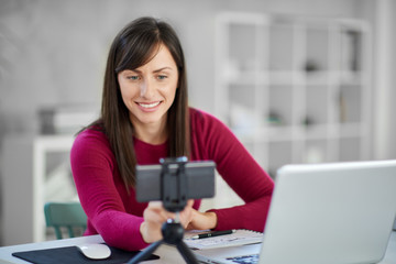 Charming Caucasian brunette dressed casual adjusting smart phone for video call. In front of her laptop, office interior.