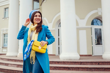 Young woman in trendy blue coat walking in city holding stylish handbag. Spring female clothes and accessories. Fashion