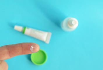 Contact lens on forefinger on light blue background.Eye care products in the background, space for text ,top view.