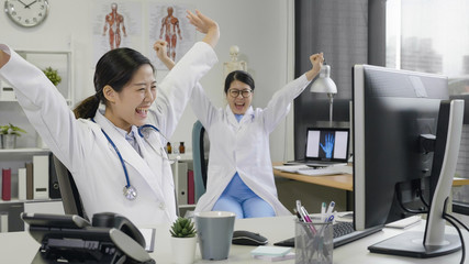 Obraz premium Happy successful female asian doctors celebrate victory in hospital office success of patient treatment. young girls medical staff workers sitting at working desk raised arms together cheerful smile