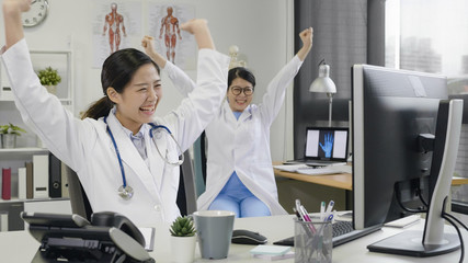 female asian doctors wearing lab coat sitting in clinic office working on computer read a good news look happy raising fist up. cheerful nurses coworker laughing about successful of patient surgery