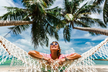 Happy smile beautiful girl in a straw hat and shorts, striped t-shirt, lying on a beach hammock between two palm trees, on the seashore of a tropical island, traveling summer vacation