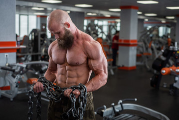 A bald, bearded bodybuilder with large relief muscles holds a heavy thick chain in his hands....