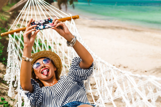 Happy Beautiful Girl In Straw Hat And Shorts, Lies On A Beach Hammock, Takes A Selfie, Between Two Palm Trees, On The Seashore Of A Tropical Island, Travels Summer Vacation