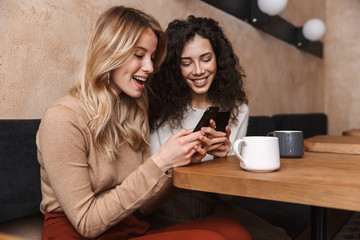 Emotional shocked girls friends sitting in cafe using mobile phone.