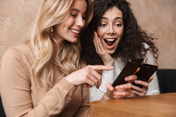 Emotional shocked girls friends sitting in cafe using mobile phone.