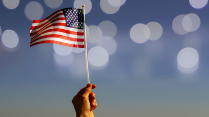 Hand holding American flag against  blue sky with sunny bokeh, vintage toned image