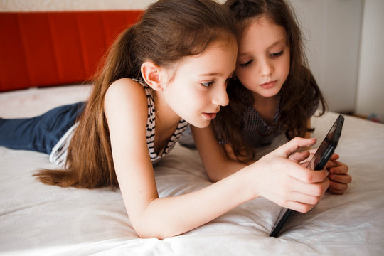 Children using digital gadgets at home. Little girls playing games on the tablet lying on the bed in the bedroom.