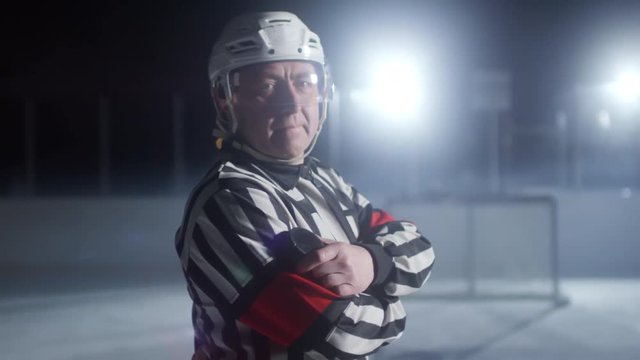 Portrait of mature male ice hockey referee in striped jersey and helmet standing with arms crossed and looking at camera