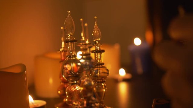 There are large/small candles and curved glass vials in the room on the table. A man holds out his hand and takes one vial. There is a mirror on the wall. Fabulous, mystical theme. Side view.