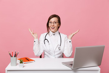 Beautiful female doctor sits at desk works on computer with medical document in hospital isolated on pastel pink wall background. Woman in medical gown glasses stethoscope. Healthcare medicine concept