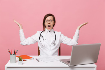 Beautiful female doctor sits at desk works on computer with medical document in hospital isolated on pastel pink wall background. Woman in medical gown glasses stethoscope. Healthcare medicine concept
