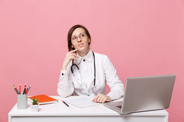 Beautiful female doctor sits at desk works on computer with medical document in hospital isolated on pastel pink wall background. Woman in medical gown glasses stethoscope. Healthcare medicine concept