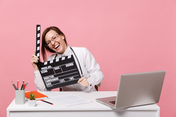 Female doctor sit at desk work on computer with medical document hold board in hospital isolated on pastel pink wall background. Woman in medical gown glasses stethoscope. Healthcare medicine concept.
