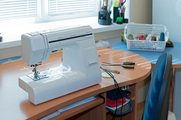 wooden table with white sewing machine, blue chair, spools of thread in basket near window in sewing studio