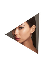 Cropped half-turn geometric portrait of woman with black flicks. The girl is wearing silver earring with round pendant, adorned with long pendants looking at camera behind triangle-shaped foreground.