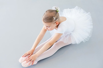Cute little ballerina is doing stretching exercises on light background. Child girl is studying in...