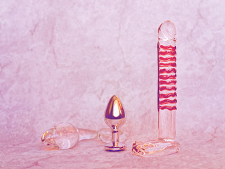 Three different sex toys (glass dildo, glass and metal butt plug) are on a light gray background...