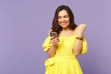 Smiling young woman in summer dress using mobile phone, typing sms message, doing winner gesture isolated on pastel violet background. People sincere emotions, lifestyle concept. Mock up copy space.