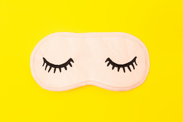 Pastel pink sleep mask with closed eyes embroidered on it with eyelashes on bright yellow neon paper background. Top view, flat lay. Concept of vivid dreams. Accessories for girls and young women.