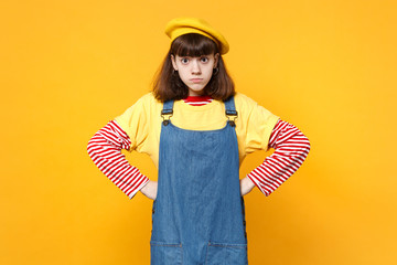 Confused puzzled girl teenager in french beret, denim sundress standing with arms akimbo isolated on yellow wall background in studio. People sincere emotions, lifestyle concept. Mock up copy space.