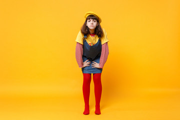 Full length portrait of dissatisfied girl teenager in french beret denim sundress standing isolated on yellow wall background in studio. People sincere emotions, lifestyle concept. Mock up copy space.